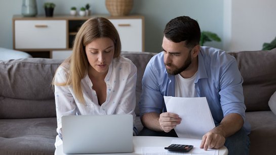 Ein Paar sitzt mit Laptop und Schreibblock auf dem Sofa und kalkuliert