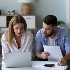 Ein Paar sitzt mit Laptop und Schreibblock auf dem Sofa und kalkuliert