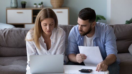 Ein Paar sitzt mit Laptop und Schreibblock auf dem Sofa und kalkuliert